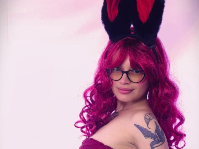 KoraSweet with pink curly hair, black glasses, and bunny ears, smiling at the camera, showing a tattoo on her right shoulder, wearing a strapless purple top.