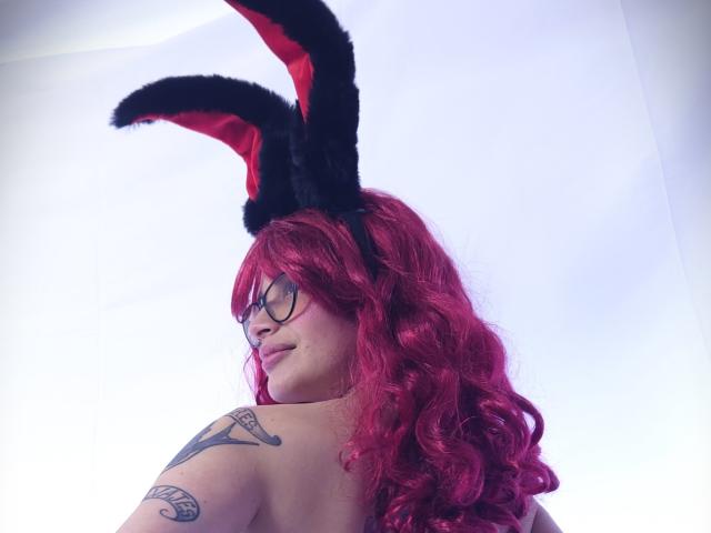 KoraSweet with red curly hair, black bunny ears, glasses, tattoo on shoulder, looking back, showing bare back. Her tits are not visible. She's cute and sexy.
