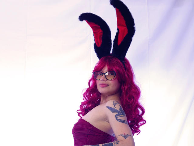 KoraSweet with red curly hair, black furry bunny ears, glasses, and tattoos, wearing a strapless maroon top, showing her ass, and a cute smile.