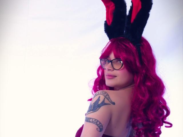 KoraSweet with red wavy hair, black fuzzy bunny ears, glasses, and a tattoo on her right shoulder, looking over her shoulder, showing her bare back and butt.