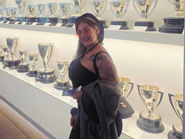 AzhunaStar stands in a trophy display room. She's wearing a black dress, a black beret, and red-tinted glasses. Her right arm, covered in a detailed tattoo, is draped with a dark gray jacket. She holds a black clutch. The room is filled with shiny silver trophies on a white shelf. Her long brown hair is partially visible under the beret. She smiles at the camera.