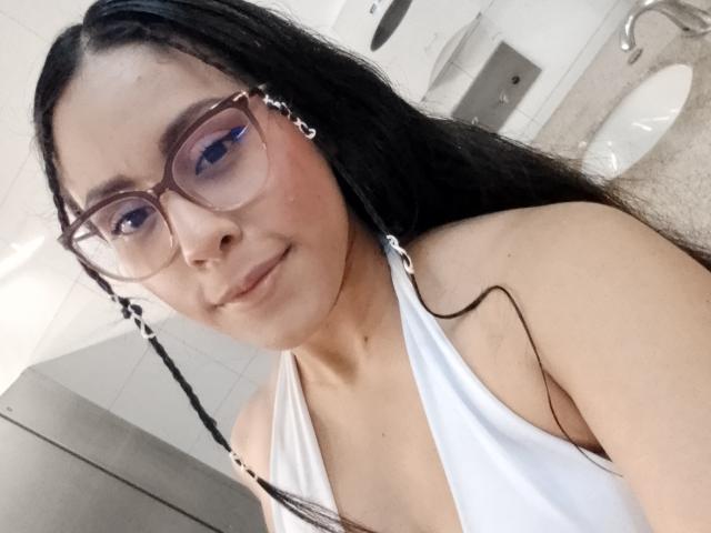 Close-up of MarilynHills with long black hair in a side braid, wearing large brown-rimmed glasses and a white halter top. She has fair skin, light pink eyeshadow, and a subtle smile. The white tiled bathroom is in the background with a sink and faucet visible. Her face is slightly angled to the right, and the photo has a casual, slightly blurry quality. Her glasses have a metallic sheen, and her hair is smooth with a slight wave.