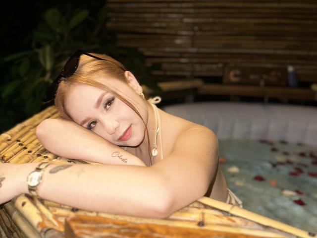 SofiaDelRey in a bamboo hot tub, arms resting on the edge, smiling, blonde hair, pink lipstick, tattoo on arm, wristwatch, sunglasses on head, water with petals.