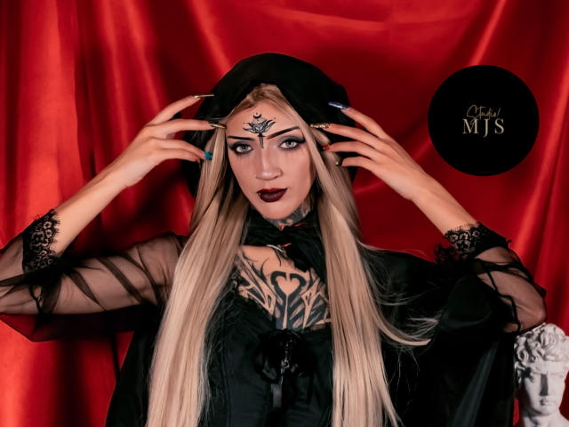 AmellieJones with long blonde hair, black sheer dress, dark lipstick, tattooed chest, black headscarf, and detailed forehead tattoo, holding her headscarf, red background, and black logo.