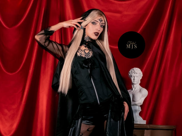 AmellieJones with long blonde hair, black gothic dress, and sheer black robe, exposing black lace and tattooed chest, standing in front of a white bust on red velvet.