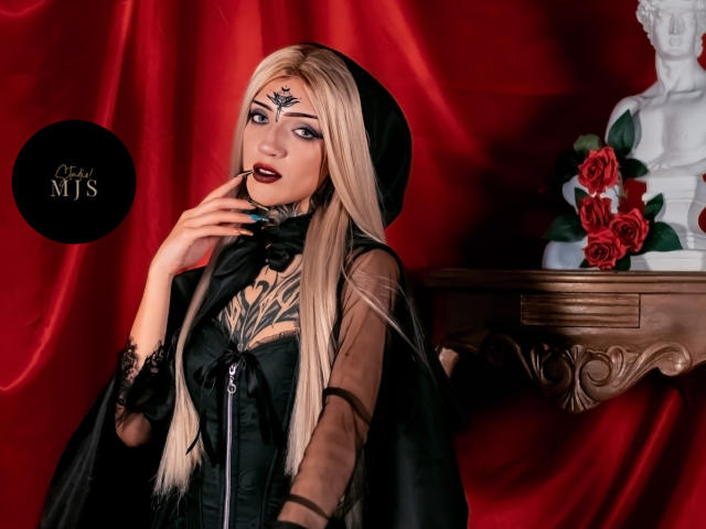 AmellieJones with blonde hair, black goth dress, and tattoos, holding a cigarette, red velvet background, black cape, white bust with red roses, dark lipstick, and a black hat.