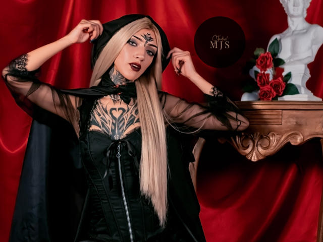 AmellieJones with long blonde hair, black gothic dress, sheer sleeves, tattoos on chest, dark lipstick, holding black hood, red background, white statue, red roses, wooden shelf.