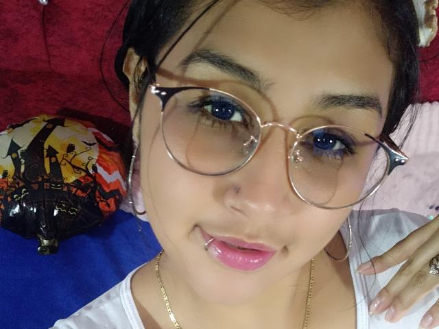EmiilyJohamson with glasses, blue eye makeup, pink lipstick, gold necklace, and tongue out, lying on red and blue fabric, with a colorful bag on the left.