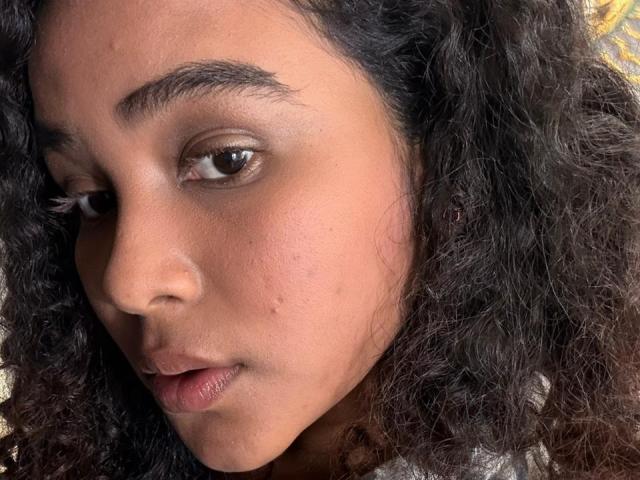 AnnyRestrepo's face, showing dark curly hair, brown eyes, and a slightly open mouth with a small pimple on her left cheek. She's wearing a subtle, dark brown eyeshadow.