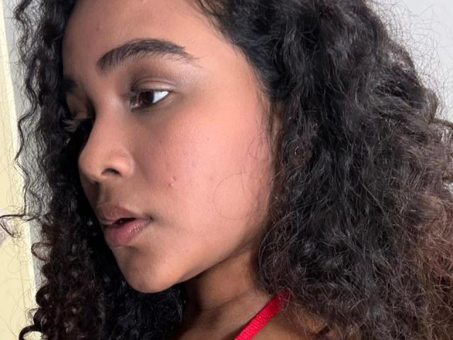 Close-up of AnnyRestrepo's face, showing a side profile. She has dark curly hair, brown eyes, and a slightly open mouth. Her skin is a medium brown tone with visible pores and a few small blemishes. She's wearing a red necklace. The lighting is soft, highlighting her facial features. The texture of her hair is curly and dense, contrasting with the smoothness of her skin. The composition focuses on her profile, emphasizing her natural beauty and subtle expression.