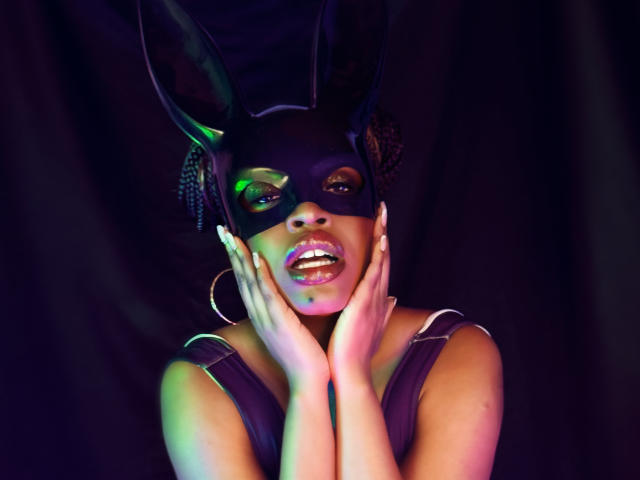 NatashaSparks with dark mask, purple dress, hoop earrings, hands on face, green and purple lights on skin, looking seductive, tall black bunny ears on head, lips parted slightly.