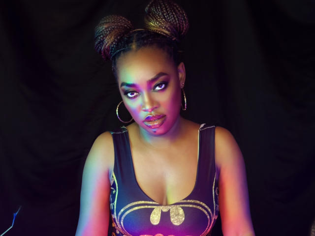 NatashaSparks has dark skin, big boobs, and braided hair in two buns. She's wearing a black dress with gold accents and large hoop earrings. Glowing purple and blue lights.