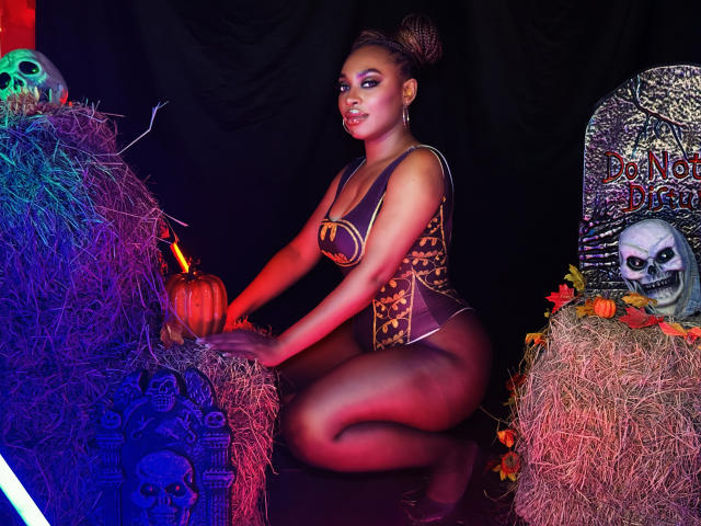 NatashaSparks kneeling, wearing a Halloween-themed corset, her brown skin and curly updo, with colorful lights, hay bales, a pumpkin, skull decor, and a spooky gravestone. Boobs, ass.