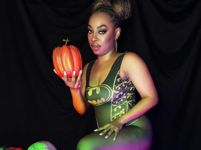 NatashaSparks with big boobs in a green, gold-embroidered bodysuit, holding a glowing pumpkin, dark curly hair in a bun, wearing hoop earrings, looking at the camera.