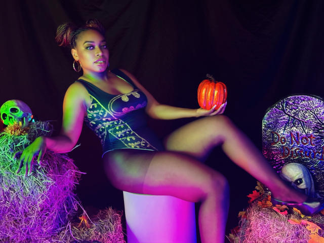 NatashaSparks with brown skin, curly red hair, wearing a Batman-themed bodysuit, holding a pumpkin, sitting on a white cube, surrounded by hay, skull, and "NO" sign. Boobs, ass, legs.