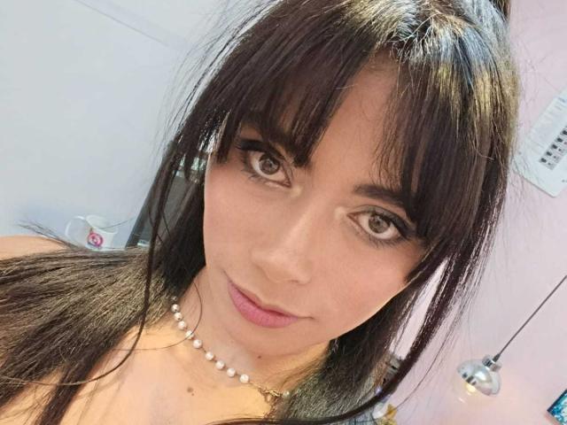 AnnaMooree tem cabelo castanho escuro, franja e um colar de pérolas. Ela está olhando para a câmera com olhos castanhos, lábios rosados e um leve sorriso. Seus seios estão parcialmente visíveis..
