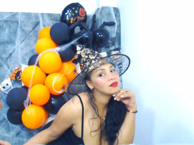 MelanyContrera with dark hair, wearing a black witch hat, red lipstick, and a black dress, sitting next to orange and black balloons, looking up with a slight smile.