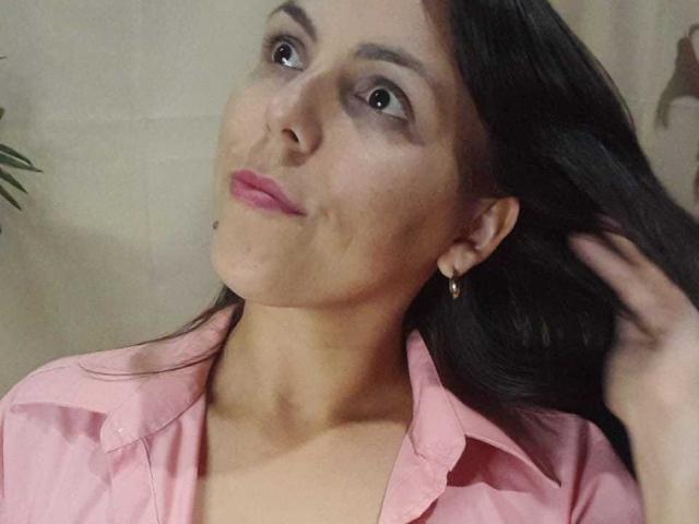 AfroditSexx has brown eyes, pink lips, and long dark hair, wearing a pink shirt, with a hand in her hair, and small gold earrings, looking up with a slight pout.