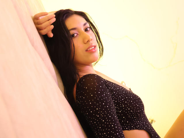 SaraRousse with long black hair, wearing a black, sheer, polka-dotted top, leaning against a wall, smiling, looking at the camera, right arm raised, glowing yellow background.