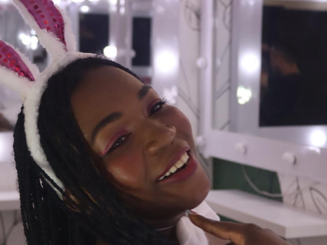 TyraFox69 with dark skin, pink eyeshadow, white bunny ears, and black braids, smiling with her head tilted. She's wearing a white shirt, and her hand is near her neck.
