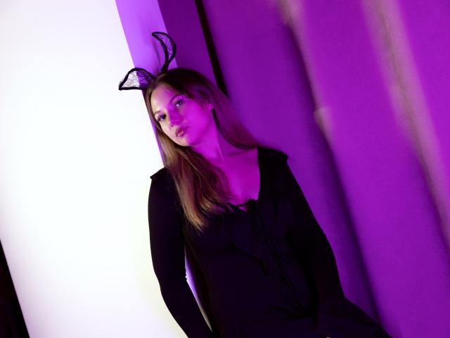 MariaRobinson with long brown hair, wearing a black dress and black lace bunny ears, leaning against a purple-lit wall, looking to the side with a slight smile.