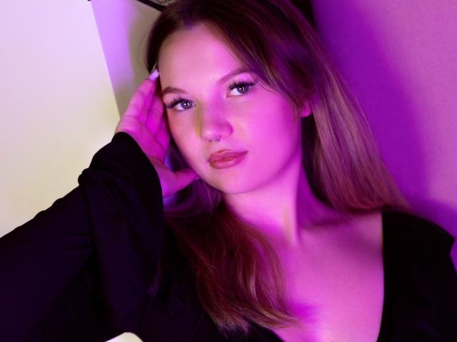 MariaRobinson with pinkish-purple lighting, black top, nose piercing, long brown hair, hand on cheek, looking at camera, slightly parted lips, blue eyes, soft makeup, subtle expression.