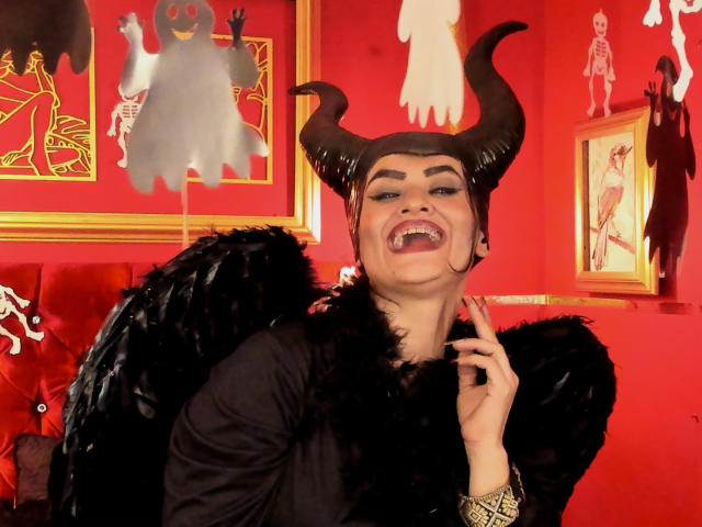 Anissa Ali con corna da diavolo, ali nere piumate e pelliccia, che sorride mostrando i canini, indossa un abito nero, sfondo rosso e un'opera d'arte spettrale incorniciata. La sua mano è vicino al mento..