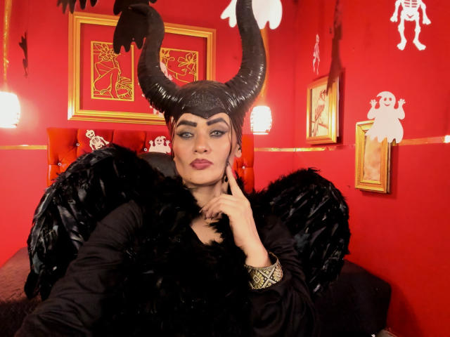 AnissaAli with black demon horns, black feather wings, and black fur dress, looking at camera with finger on chin, red wall, gold frames, and white ghost cutouts in background.