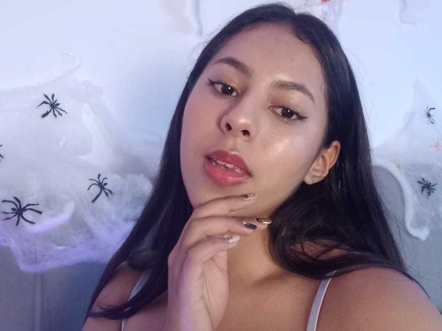 SoffiRousee has shiny skin, black hair, and pink lips, with her hand near her face, showing gold and black nail polish. She's wearing a white top.