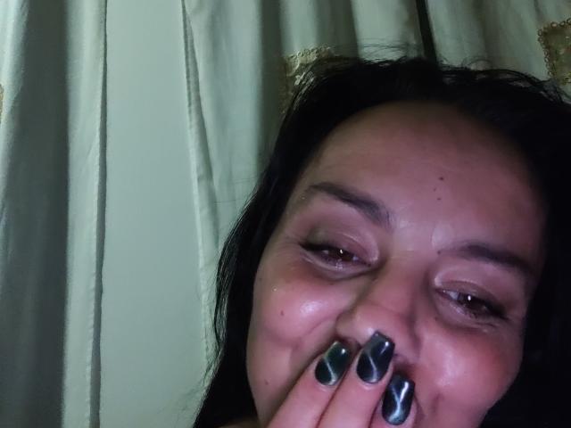 AnaPiyas with dark hair, brown eyes, and shiny black nails, touching her nose, smiling slightly, with a subtle sheen on her face, in a dimly lit room.
