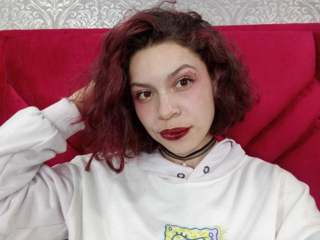 SophieBlake with red lipstick, curly dark red hair, white hoodie, black choker, and pink eyeliner, resting hand on head, looking at camera, slight smile.