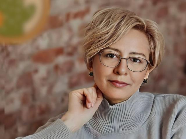 VeloriaSelena with short blonde hair, glasses, and a grey turtleneck, smiling softly, hand near chin, wearing black earrings, looking cute and smart.