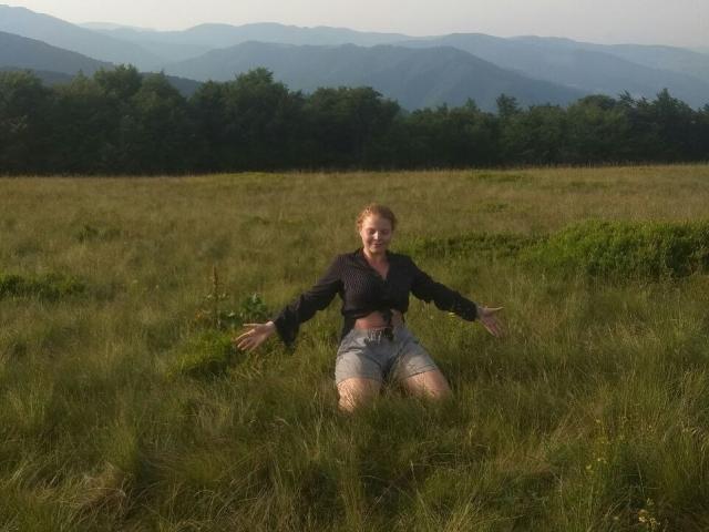 RenesmeeSunset, with her tits visible under a black shirt, is kneeling in tall grass with arms out, wearing gray shorts, in a field with mountains in the background.