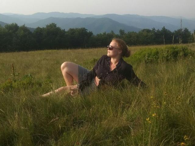 RenesmeeSunset with tits covered, sitting in tall grass, wearing glasses and a dark shirt, legs bent, looking up, in a sunlit meadow with mountains and trees in background.