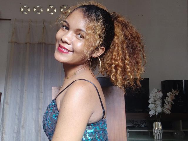 NazziaSexi with curly brown hair in a ponytail, smiling with red lipstick, wearing a blue sequined top, showing her cute ass, in a well-lit room.