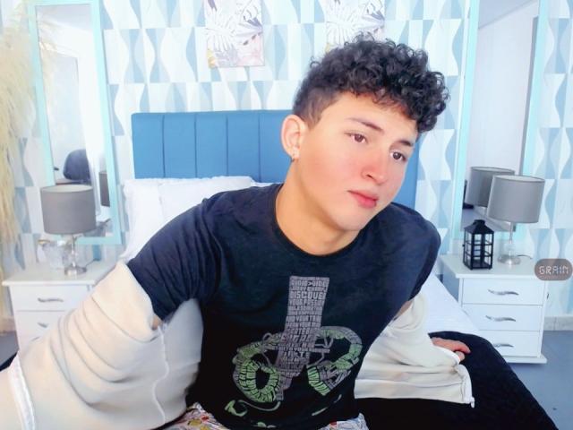 AxelSavage with curly black hair, light skin, and red lips, wearing a black shirt with a large green letter "T", sitting on a bed with white pillows.