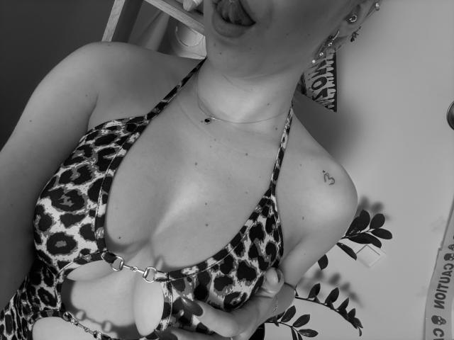 Black and white photo of SugarrX with leopard print bikini, showing cleavage, tongue out, small heart tattoo on shoulder, earrings, and necklace, holding a leafy plant.