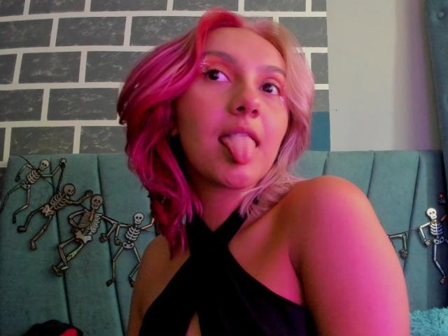 AlterEgoSecond with pink and blonde hair, black criss-cross top, tongue out, facing camera, showing off bare shoulders, colorful lighting, and skeleton decorations on teal headboard.