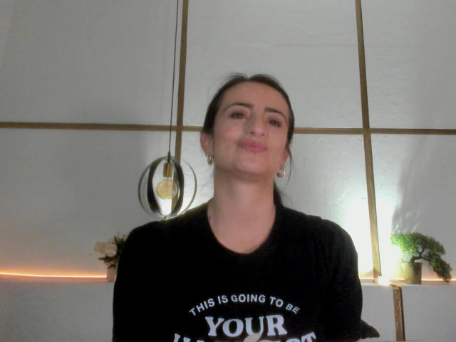 BrendaCantorel, puffy lips, black shirt with white text, brown hair in a ponytail, earrings, white ceiling with gold lines, hanging light, slight smile, standing, medium shot.