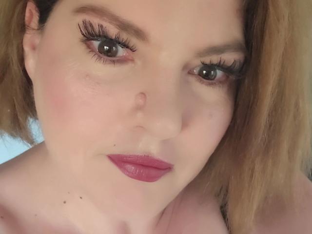 Le visage de Joyfull-hot, peau pâle, yeux marron, longues cils, rouge à lèvres rose, légère rougeur sur les joues, cheveux blonds, petite tache de beauté discrète sous le nez, blush rose clair, léger sourire, pas de vêtements visibles..