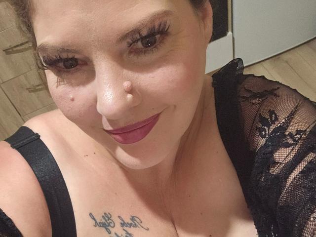 Joyfull-hot, eine Frau mit heller Haut, braunen Augen und rotem Lippenstift, trägt schwarze Spitze, zeigt ihre Brüste, hat ein Tattoo mit der Aufschrift "Live love laugh" und einen niedlichen Nasenring..