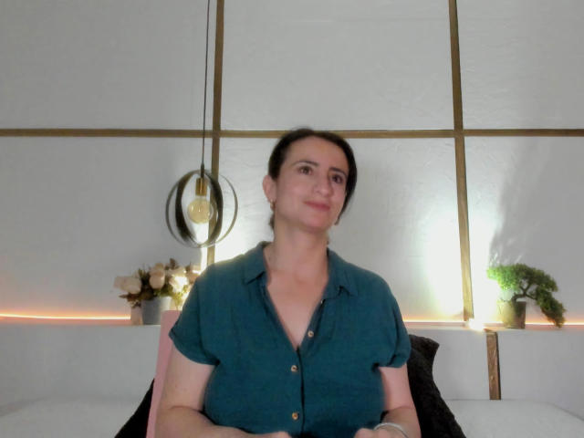 BrendaCantorel, chubby with small tits, wearing a teal button-up shirt, sits on a pink chair, smiling slightly, dark hair in a messy bun, with a potted plant behind.