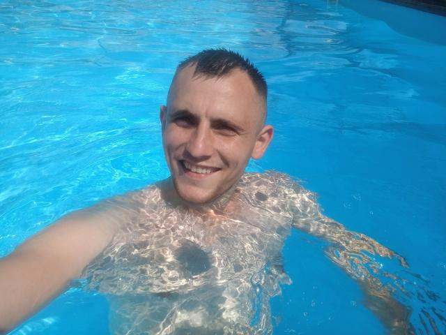 SmileGraham, shirtless, with a grin, short dark hair, and a small goatee, in a blue pool, his chest and nipples slightly visible through the water.