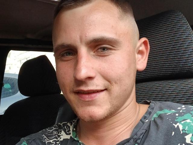 SmileGraham has a shaved head, light green eyes, and a small goatee. He's wearing a gray camo shirt, sitting in a car with black striped seats.