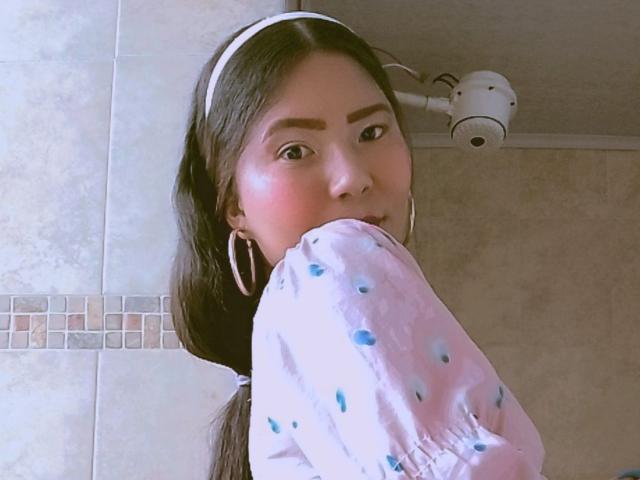 JadeFoxx porte une chemise rose à pois, de grandes boucles d'oreilles, un bandeau blanc et a de longs cheveux bruns. Elle regarde par-dessus son épaule, avec un blush sur les joues, dans une salle de bain où l'on voit un appareil photo en haut à droite..