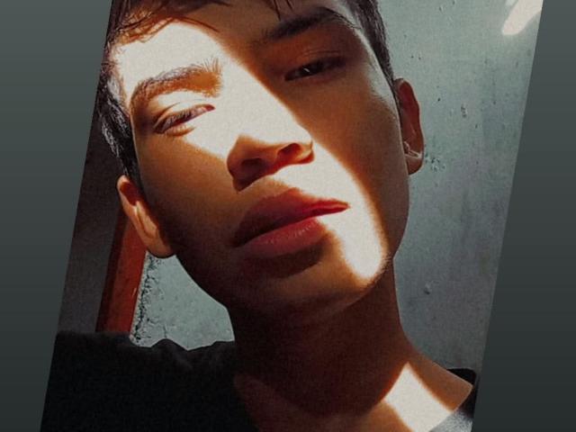 AngellDustt has a sharp face, slightly parted lips, and short brown hair, with sunlight casting a shadow over half of his face, showing his smooth, light brown skin.