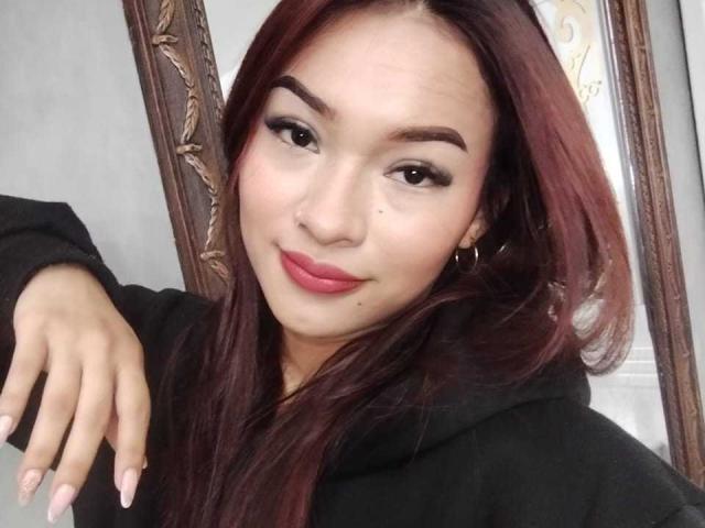 SashaHans has red hair, brown eyes, and a cute face with a small mole above her lip. She's wearing a black hoodie and has pink lipstick on.