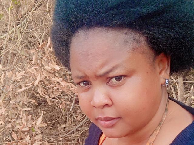 Katusi's angry face with dark green afro, brown eyes, and a small nose, wearing a dark purple top, looking to the side with a slightly pouted lower lip.
