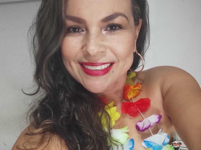 Danny69s has wavy dark brown hair, red lipstick, and a colorful flower necklace, smiling with hoop earrings, showing her tan skin and bare shoulders.