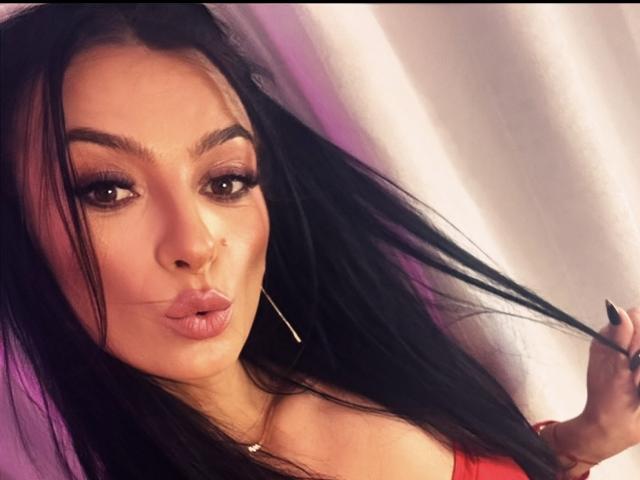 KinkyAmelie mit langen, schwarzen Haaren, vollen Lippen, die zu einem Kussmund geformt sind, braunen Augen und dunklen Augenbrauen, trägt Rot und zeigt kleine Ohrringe und einen Nasenring, mit einer Hand im Haar..